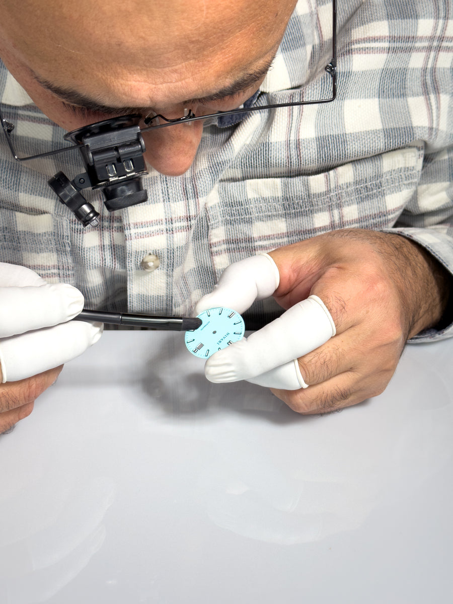 A watchmaker inspecting a dial and case under a magnifier.