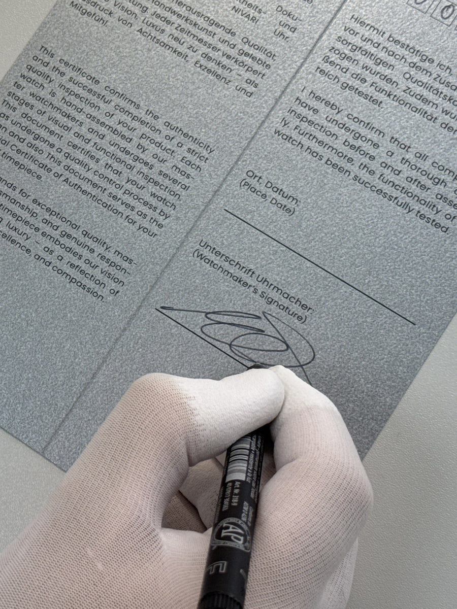 A watchmaker signing a NIVARI guarantee document beside a finished watch.